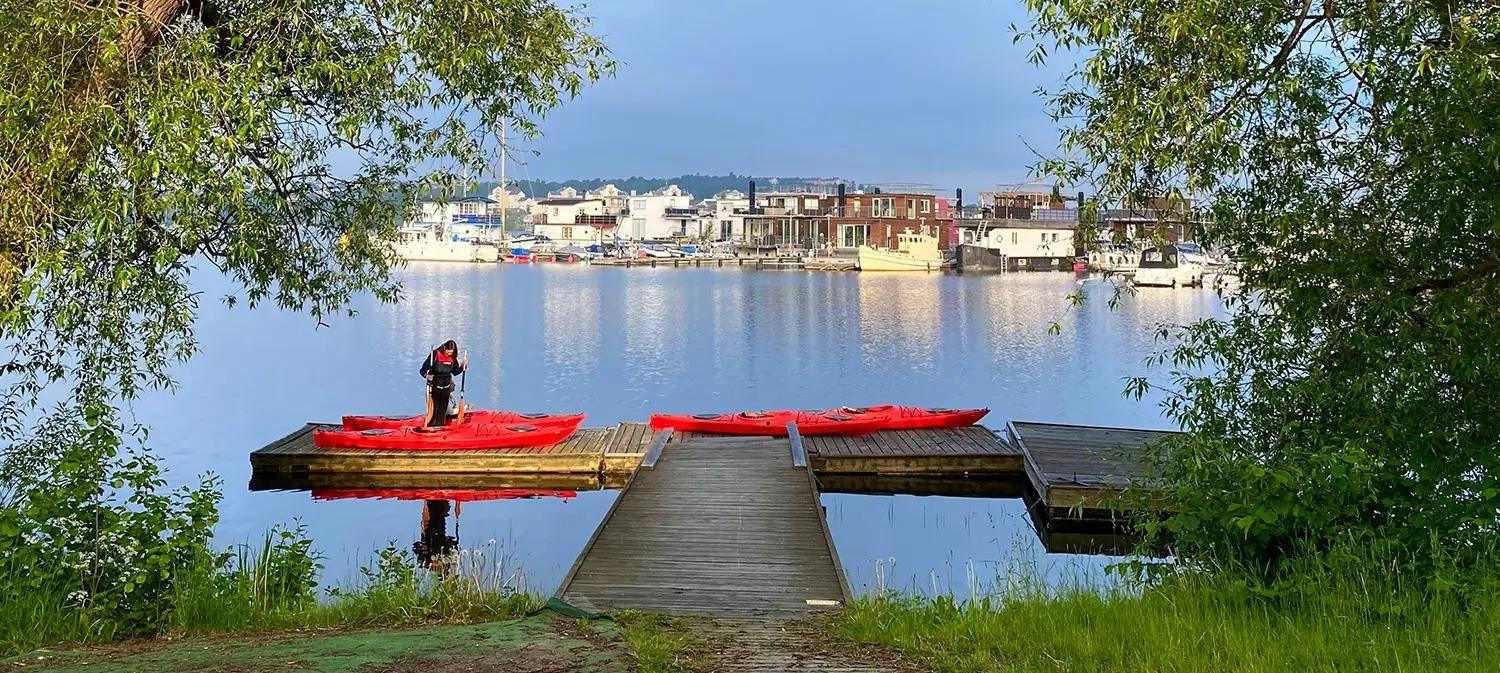 Calm, beautiful background shot of a person preparing a few red KAYAKOMAT kayaks to launch from a dock in Stockholm. Trees and calm water with city scenery in the background. 
