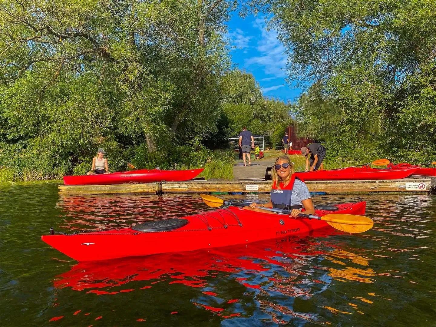 Young smiling woman paddling a red KAYAKOMAT single kayak after just launching from a dock. She's on flat calm water on a sunny day, with trees and some more people in the background preparing kayaks on the dock.  