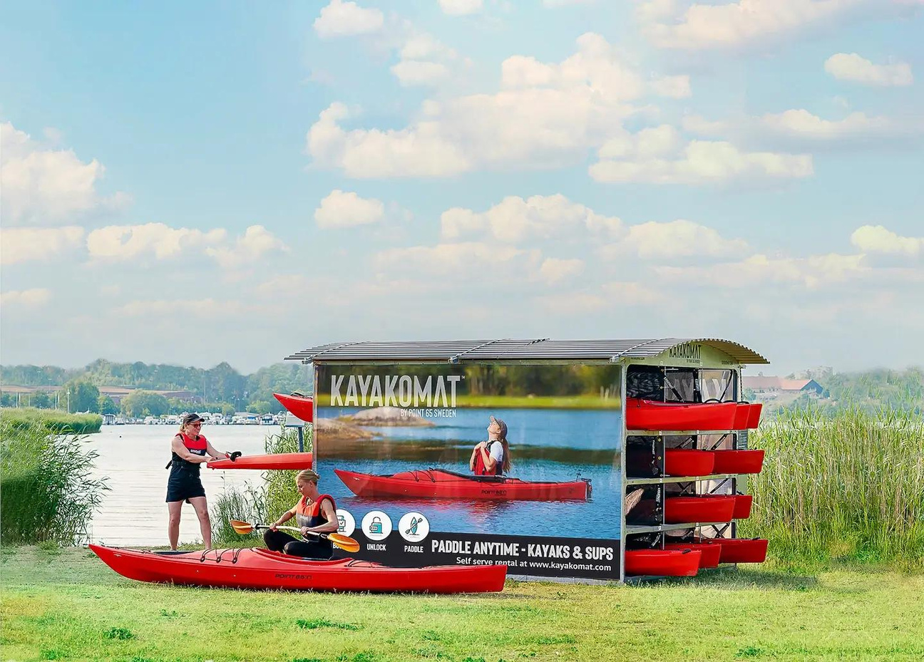 Two women preparing their red KAYAKOMAT kayaks by removing them from the self service rental station next to a calm lake on a sunny day. 
